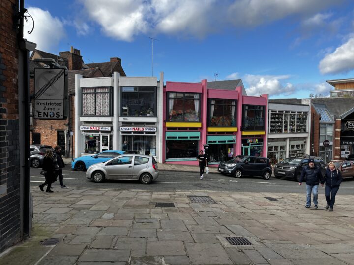 A street scene with people walking and cars parked in front of colorful buildings. The buildings include white and pink facades with large glass windows; signs read Apollo Nails Studio and Rhi Rhi. The sky is mostly blue with some clouds.