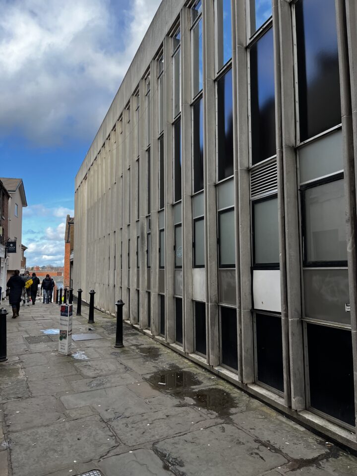 A tall, gray concrete building with narrow vertical windows lines a stone-paved sidewalk. Several people walk in the distance, and the sky above is bright blue with scattered clouds. Black bollards and a colorful pole are along the sidewalk.