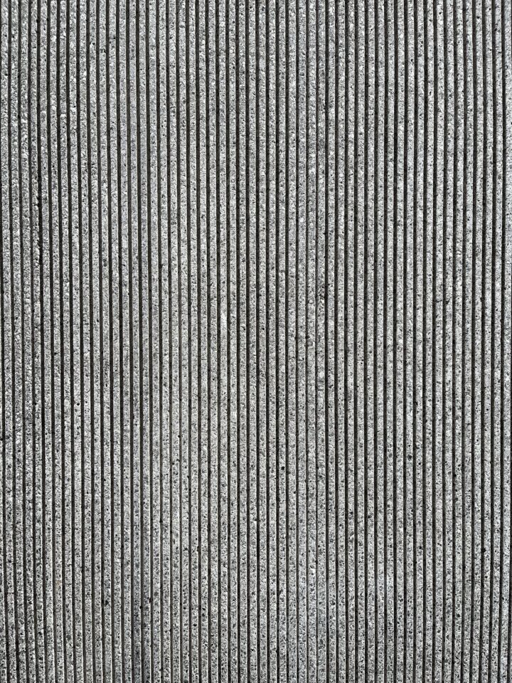 A close-up view of a textured concrete wall with evenly spaced vertical grooves. The concrete surface is rough and gray, and the grooves create a repetitive striped pattern across the image. The lighting highlights the texture and depth of the grooves.