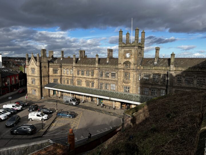 A large, historic stone railway station with a clock tower sits under a partly cloudy sky. Several chimneys and ornate details top the building. Cars are parked outside, and people walk near the entrance. A grassy embankment is visible in the foreground.