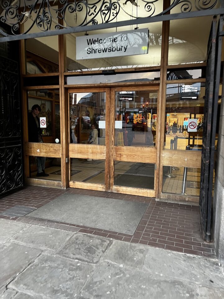Entrance to a building with glass and wooden doors, a sign above reading Welcome to Shrewsbury. People are visible inside, and outside there are stone tiles. No smoking signs are posted on the doors. Reflections show a street scene with vehicles.