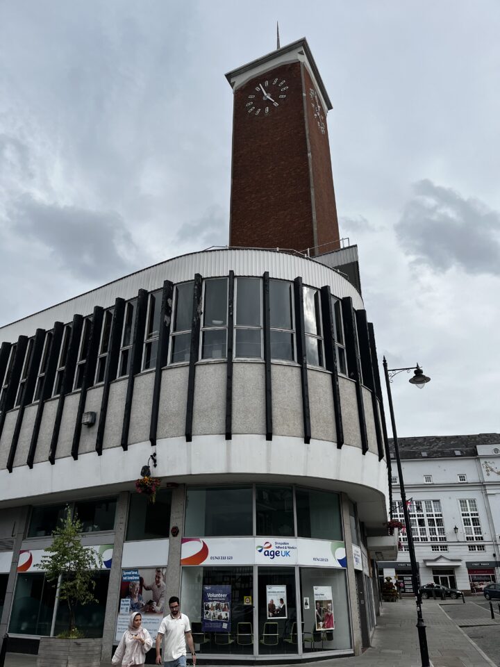 A tall red-brick clock tower rises above a modern, curved building with black vertical fins and large windows. Below, there’s an “Age UK” charity shop at street level. Two people walk past; the sky is cloudy and a streetlamp stands nearby.