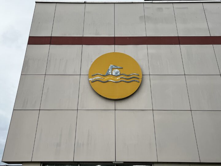 A large yellow circle sign with a swimming figure and three blue wavy lines is mounted on a beige tiled wall with a horizontal red stripe near the top. The symbol represents a swimming pool or aquatic center.
