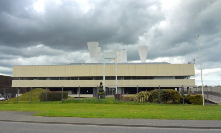 A large, modern industrial building with a flat roof and multiple white ventilation structures on top, set against a cloudy sky. The building is cream-colored with dark windows, surrounded by a fence, grass, shrubs, and a paved road in front.
