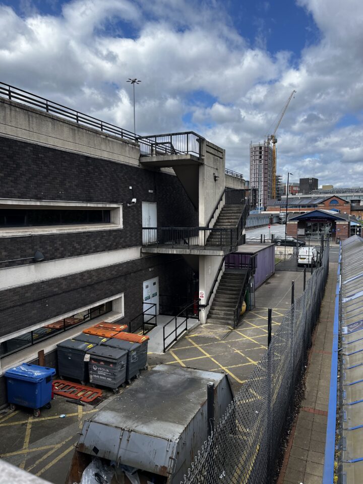 A fenced urban service yard with dumpsters and a blue bin stands beside a brick building with outdoor stairs. Yellow lines mark the ground; a van and construction crane appear in the background under a cloudy sky.