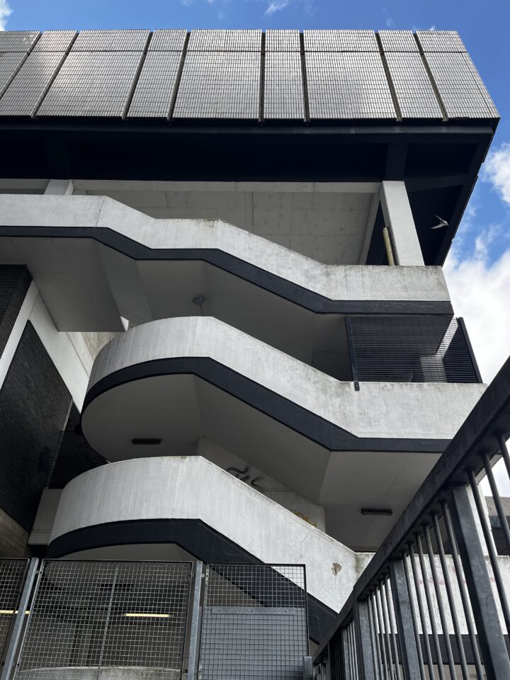 A low-angle view of a modern concrete building with three zigzagging external staircases, dark railings, and grid-like metal panels on the top level against a blue sky with scattered clouds.