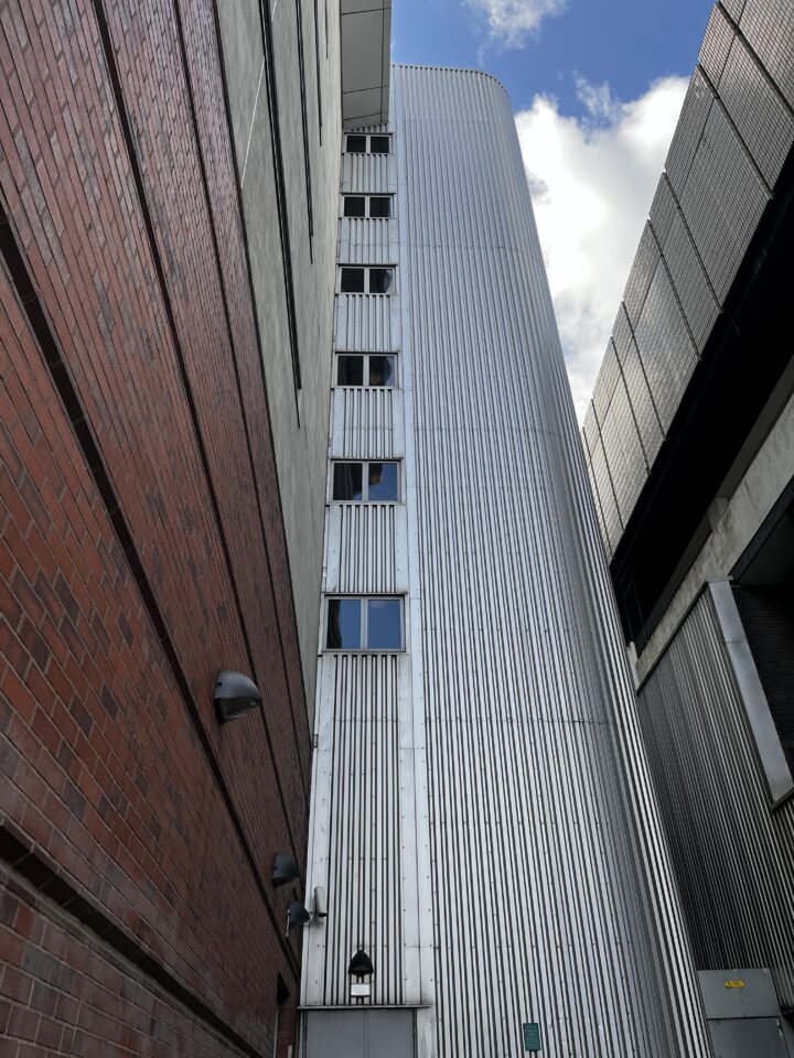 A narrow alleyway is bordered by a brick wall on the left and a tall, modern metallic building with vertical lines and narrow windows in the center. The sky with clouds is visible above, and lights and utility boxes are mounted on the walls.