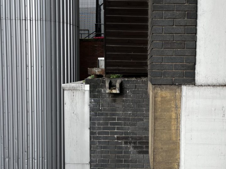 Industrial building exterior with varied textures: corrugated silver metal, dark gray and yellow brick, white concrete, and brown panels. A small concrete spout juts out, with plants growing around it. Vertical lines and contrasts dominate the image.