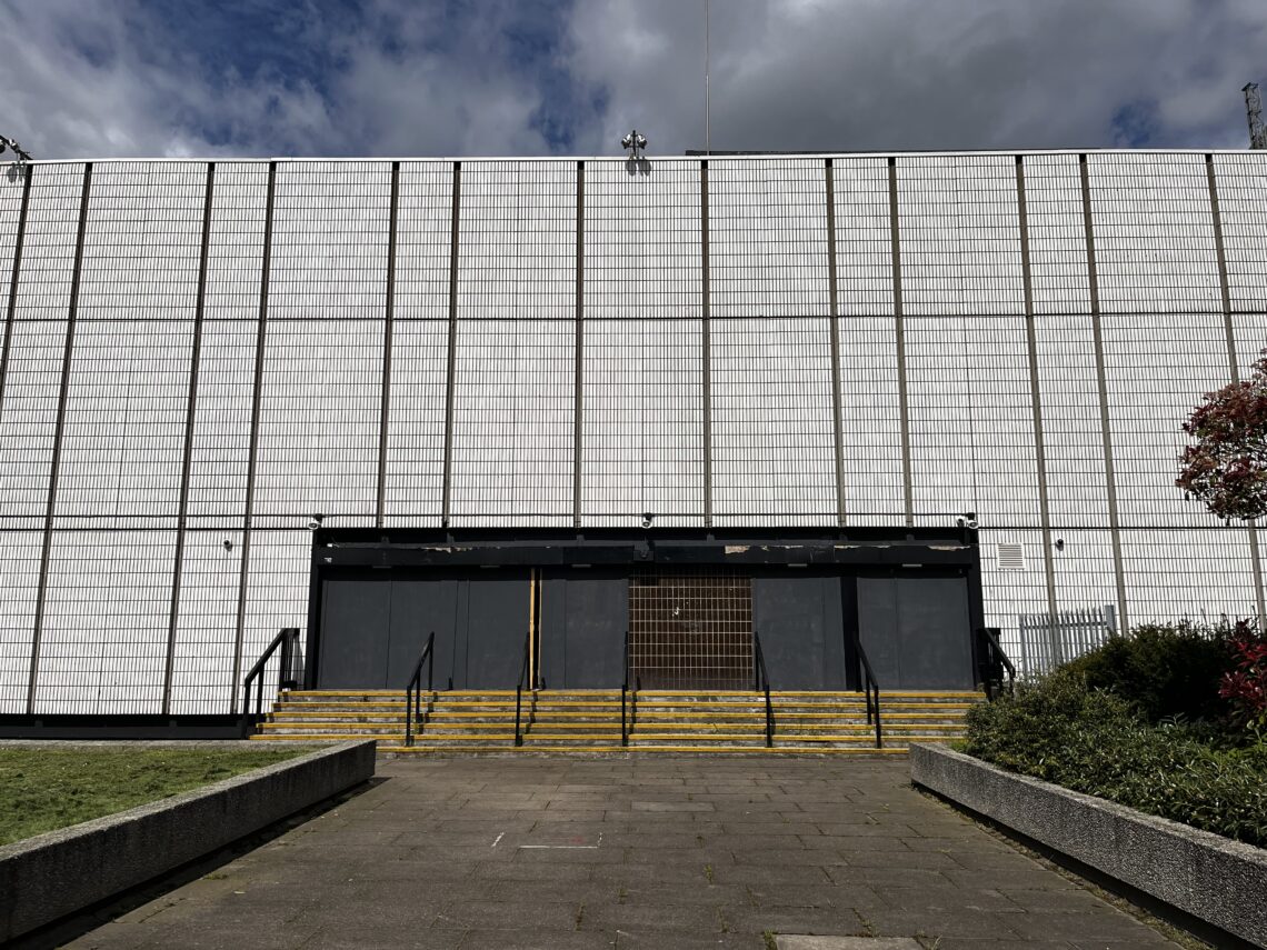 A large, windowless white building with a tiled facade, boarded-up dark doors, and yellow-striped steps. There’s a paved walkway leading up to the entrance, flanked by low concrete planters with grass and shrubs under a partly cloudy sky.