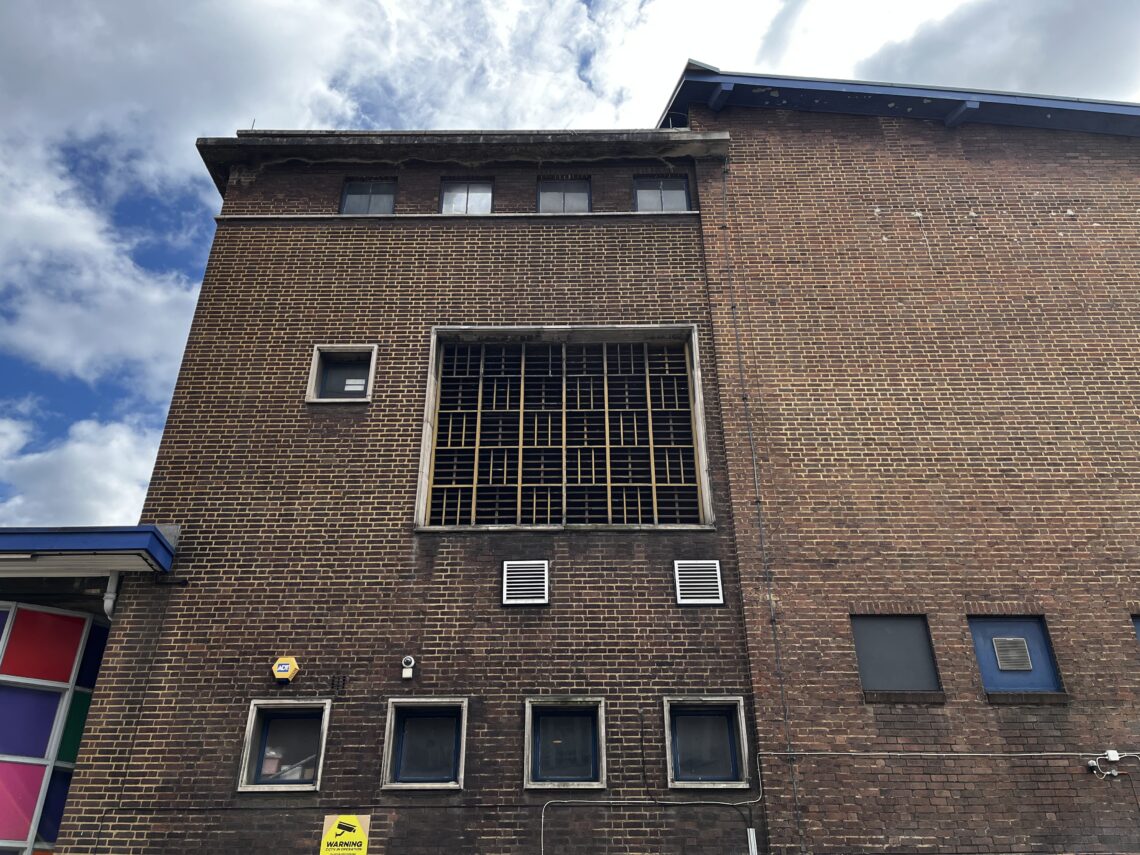 A tall brick building with a large, square window covered by a yellow grid is shown. Five smaller windows are visible below, alongside vents and security cameras. Part of a colorful structure is on the left, under a cloudy blue sky.