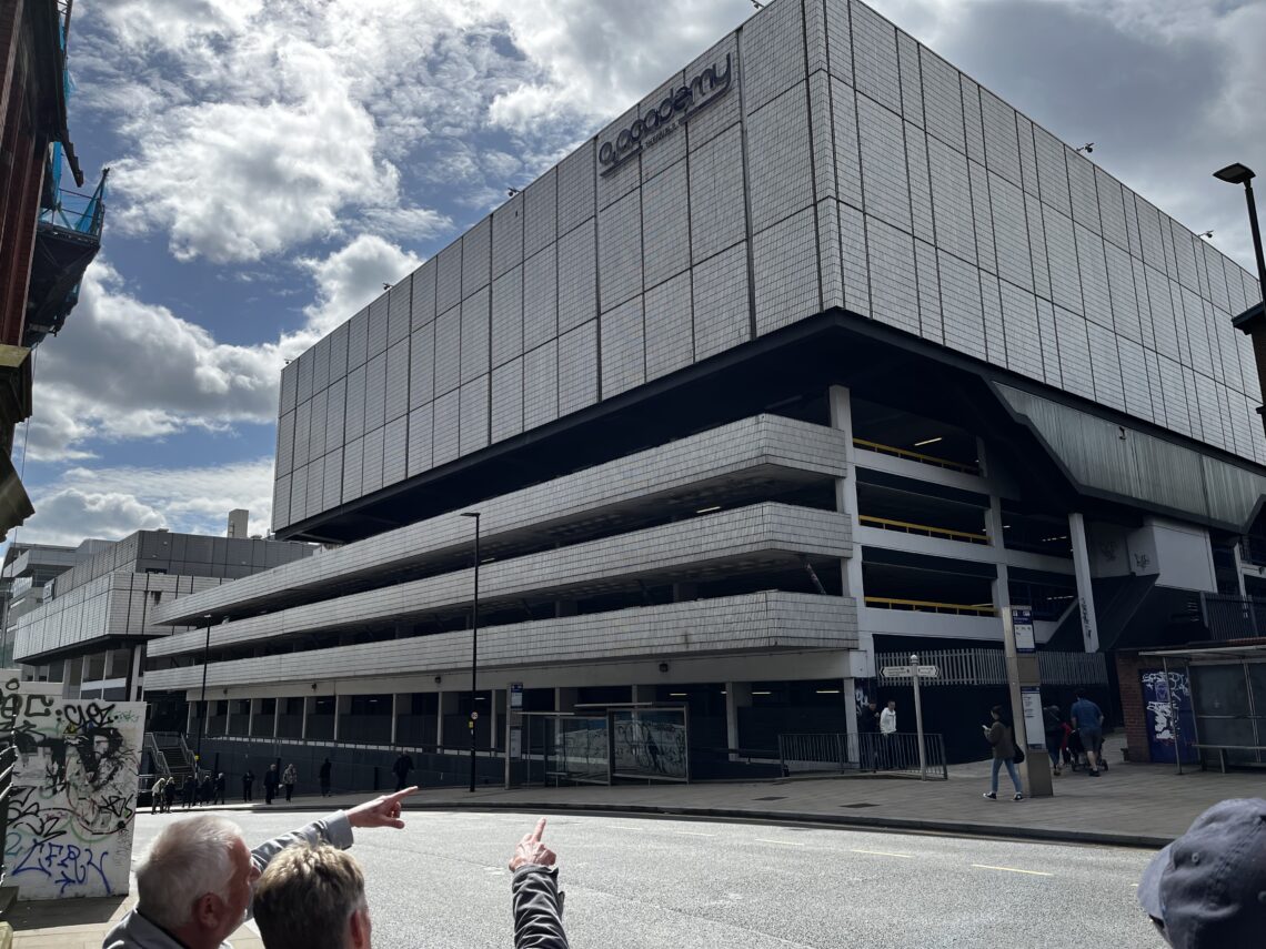 A large, modern building with a white, rectangular, tile-like facade and the sign “O2 Academy” on top. The structure includes a multi-level parking area. Several people stand and point on the street in the foreground under a partly cloudy sky.
