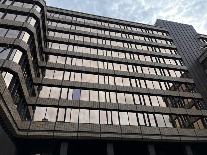 A modern office building with rectangular, reflective windows and gray tiled exterior. The structure has multiple stories and a geometric design. The sky, with light clouds, is reflected in the windows, adding a soft hue to the building’s facade.