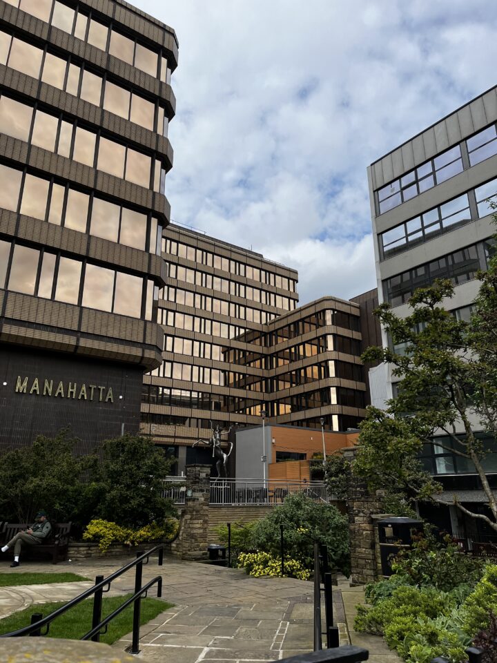 A modern urban courtyard with stone paths, flowerbeds, and benches. Surrounding the area are tall office buildings with bronze-tinted glass windows. The MANAHATTA sign is visible on one building. Trees and cloudy sky are in the background.