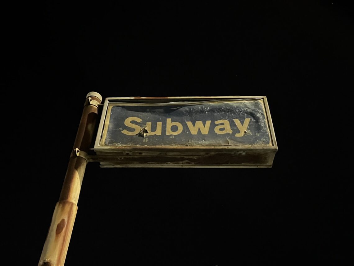 A worn rectangular sign with the word Subway in faded yellow letters stands on a rusty pole against a dark, night sky. The sign appears weathered, with chipped paint and visible marks on its surface.