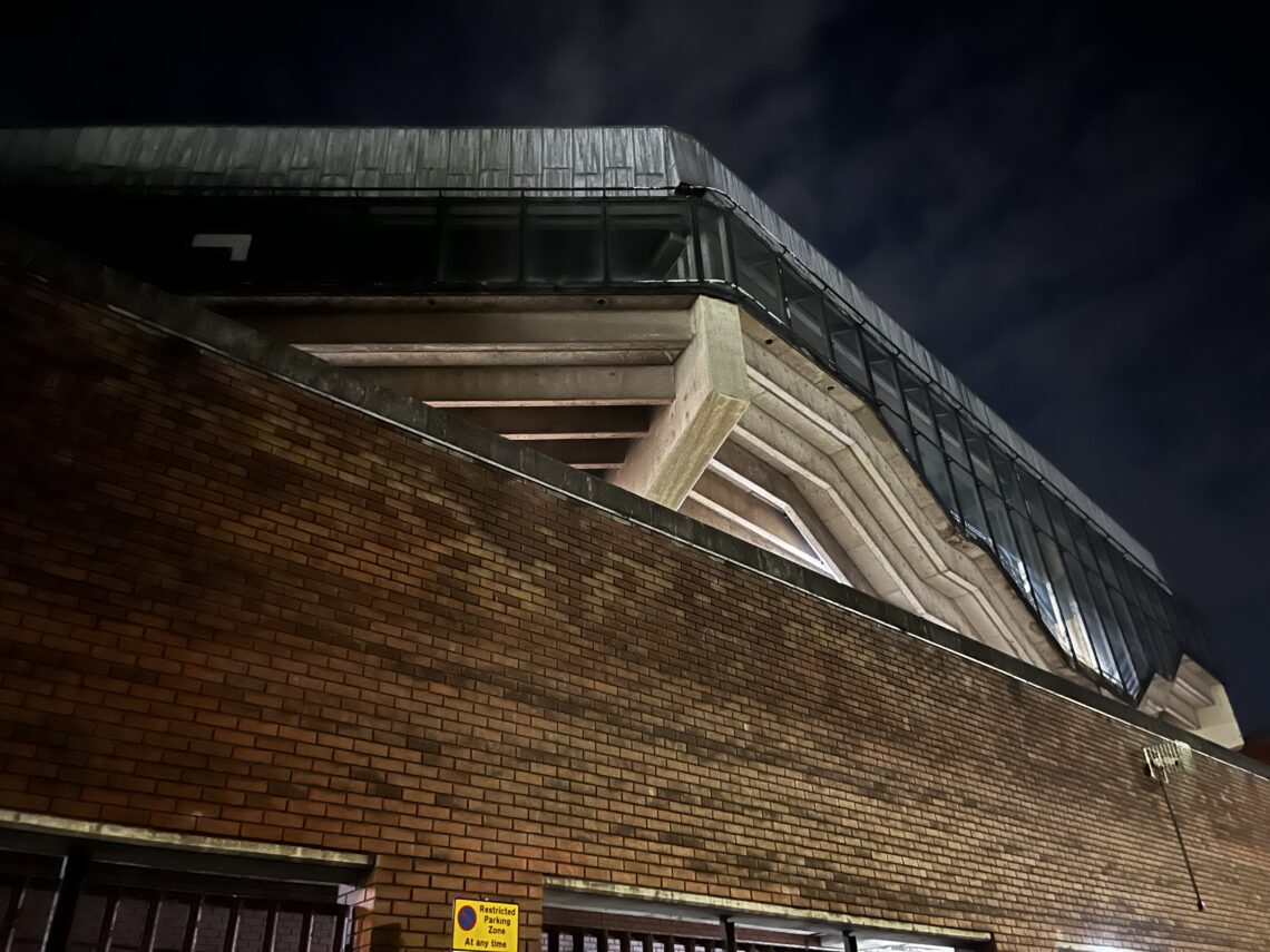 A dramatic low-angle view of a modern building at night shows angular concrete beams and large, dark reflective windows above a brick wall. The structure is illuminated by artificial light, and a small yellow sign is visible near the bottom. The sky is dark and cloudy.