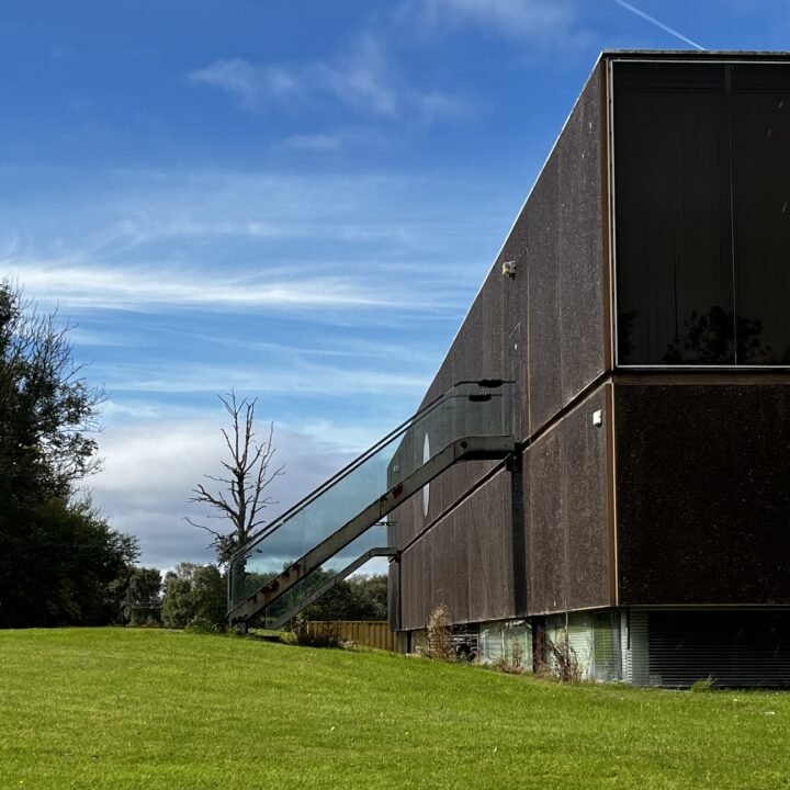 A modern building with dark, rectangular walls and large glass windows stands beside a grassy lawn. A glass-sided external staircase rises along the side. Trees, including a bare one, border the lush green lawn under a partly cloudy blue sky.