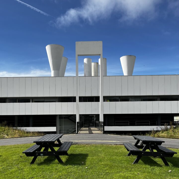 A modern white building with large, cylindrical ventilation towers on the roof stands under a blue sky. In the foreground, two black wooden picnic tables sit on a green lawn near a curved pathway leading to the building’s entrance.