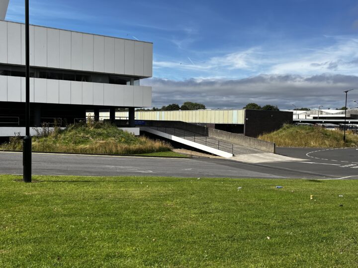 Modern white and black building with a ramp leading to an underground parking area. Green grass and weeds grow nearby. A lamppost stands on the left, and a road with roundabout markings runs in front. Sky is mostly blue with some clouds.