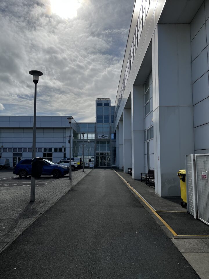 A modern, white building under a cloudy sky is shown. Cars are parked on the left, with lamp posts lining the area. A tall glass entrance is visible ahead, and a yellow recycling bin stands on the right near benches. Sunlight filters through the clouds above.
