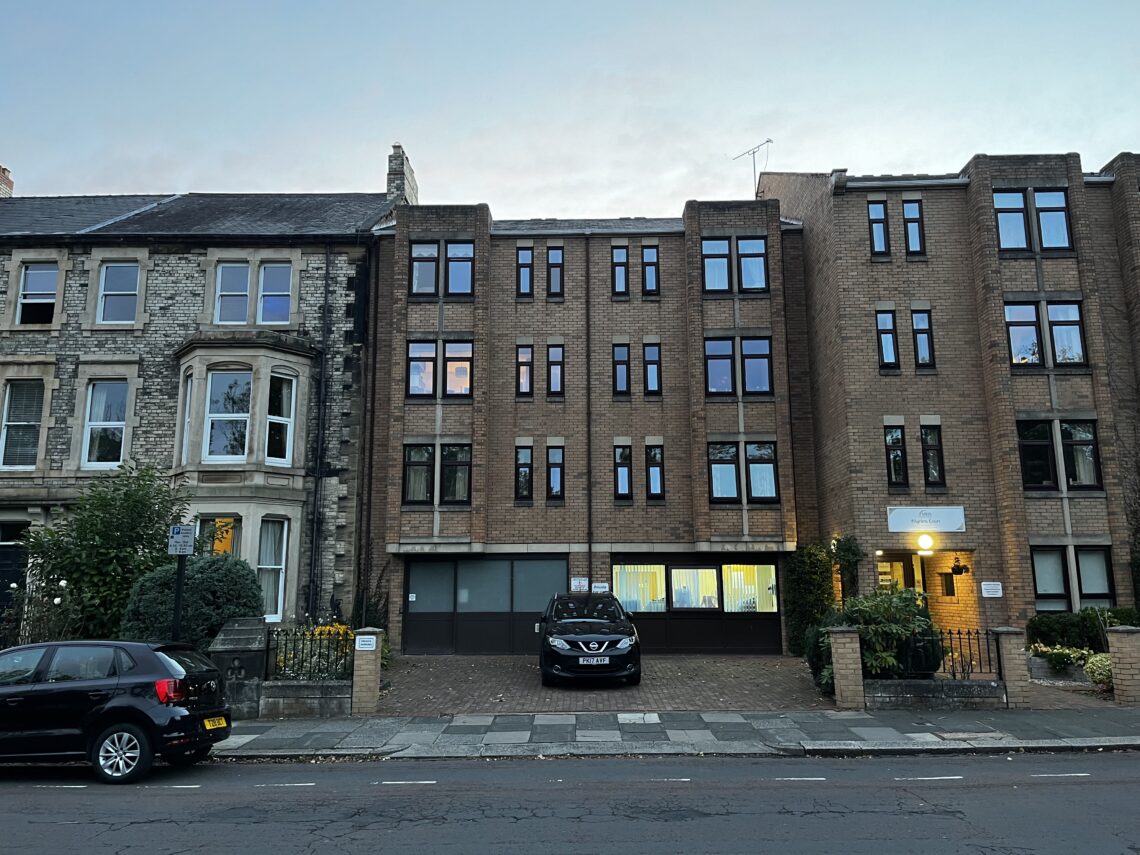 A brick and stone building with three floors and many dark-framed windows stands between older buildings. Two cars are parked in front; one is partially under the building. There’s a lit entrance on the right, and trees and plants line the sidewalk.