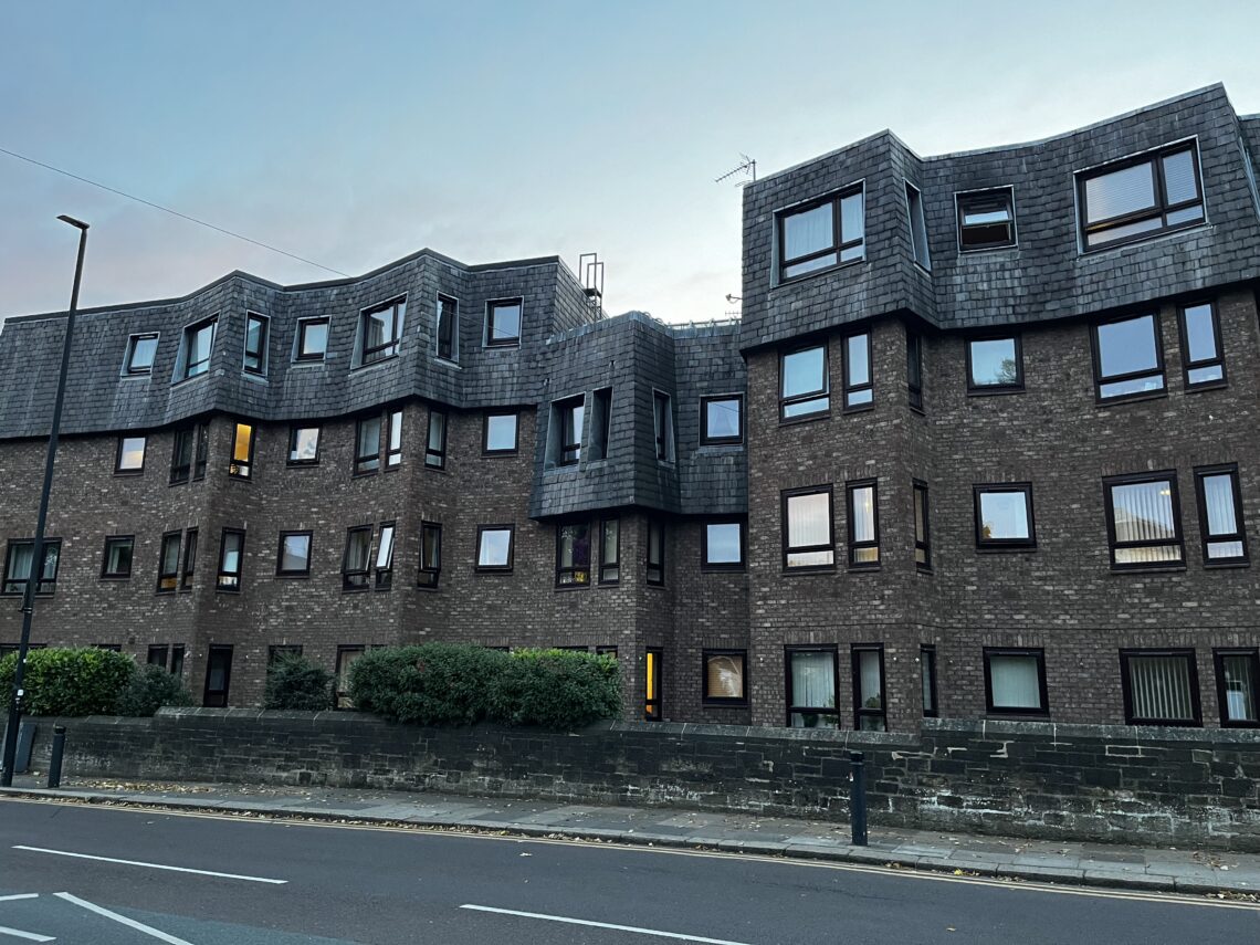 A modern, multi-story apartment building with dark brick walls and irregular, wavy rooflines. Numerous rectangular windows reflect the fading daylight. A low stone wall separates the building from a quiet street with painted road lines and a few bushes.