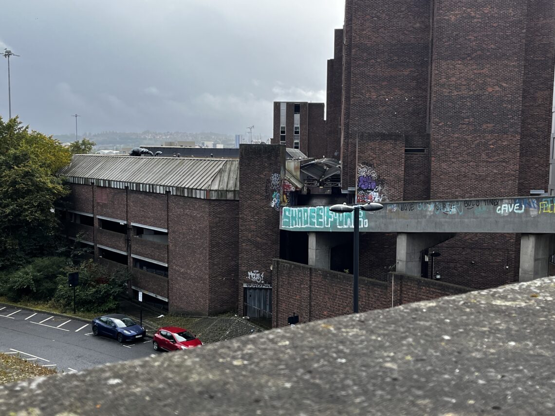 A multi-level, brutalist brick parking garage with graffiti, including a prominent “SHARE SPOTS” tag, stands next to a road with two parked cars. Trees and cloudy skies are in the background; the foreground shows a concrete barrier.