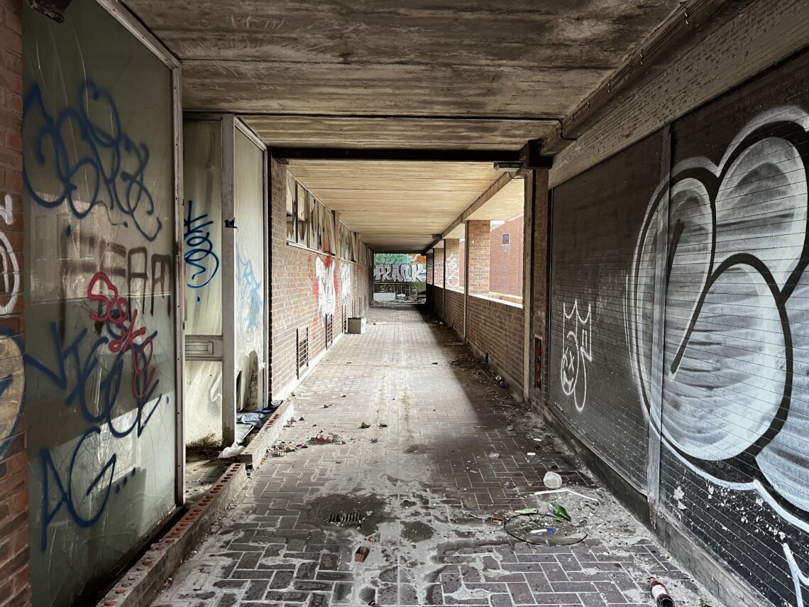A covered, abandoned walkway with brick walls and a tiled floor is littered with trash and debris. Graffiti covers the walls on both sides, and a large, colorful tag is visible at the far end. Natural light filters in from the open front.