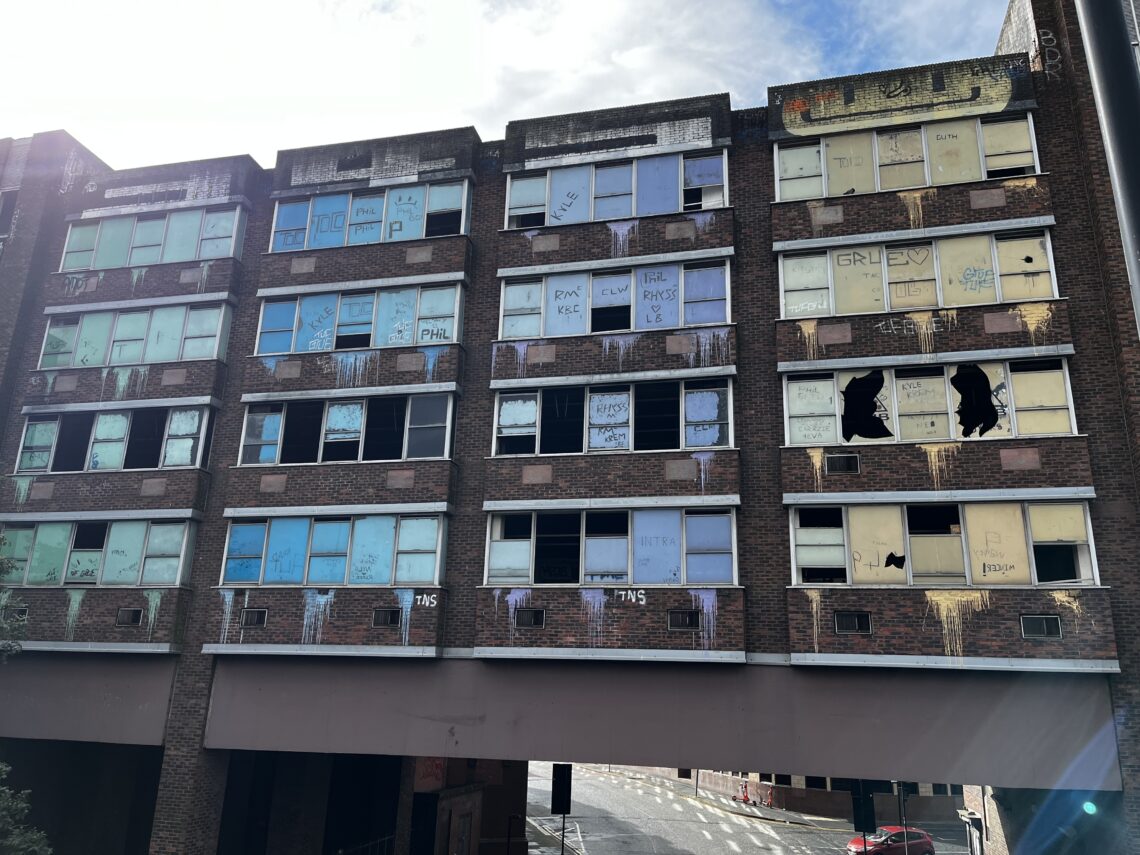 A large, abandoned brick building with six floors has many broken and boarded-up windows, most covered with faded blue, yellow, and white boards covered in graffiti. Sunlight partially shines on the building. A street runs beneath an overpass at ground level.