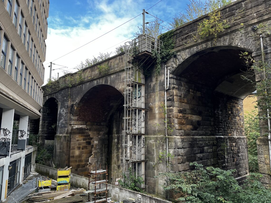 A stone railway bridge with two large arches stands beside a modern building. Metal scaffolding is attached to the bridge. Vines grow on the stonework, and construction materials are stacked below. Graffiti is visible on both the bridge and the nearby building.