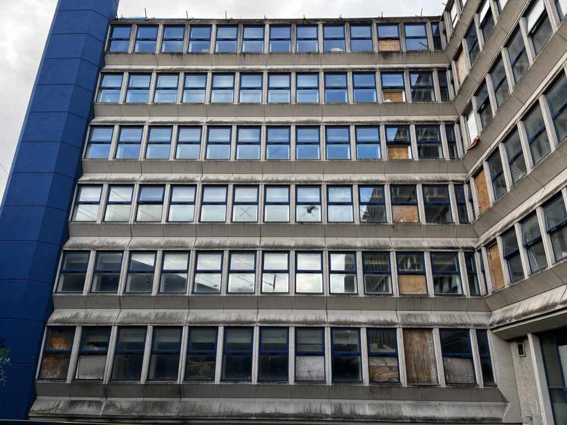 A large, rectangular, concrete office building with many rows of blue-framed windows. Some windows are boarded up with wood, and there are visible stains on the building’s surface, suggesting age or neglect. The sky is cloudy.