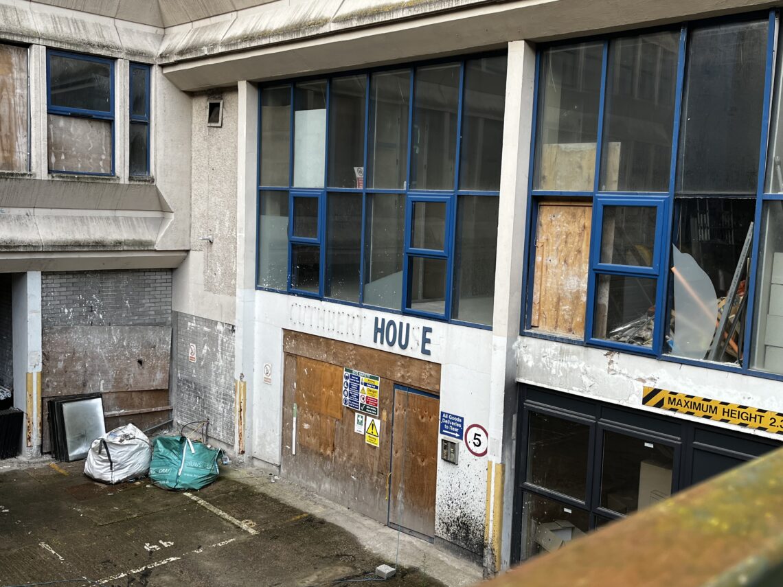 A rundown building with boarded-up windows, blue window frames, and signs of neglect. The sign reads HOUSE with missing letters. Warning signs and a speed limit sign are on the wall. Debris, green sacks, and a broken mirror are visible in the dirty courtyard.