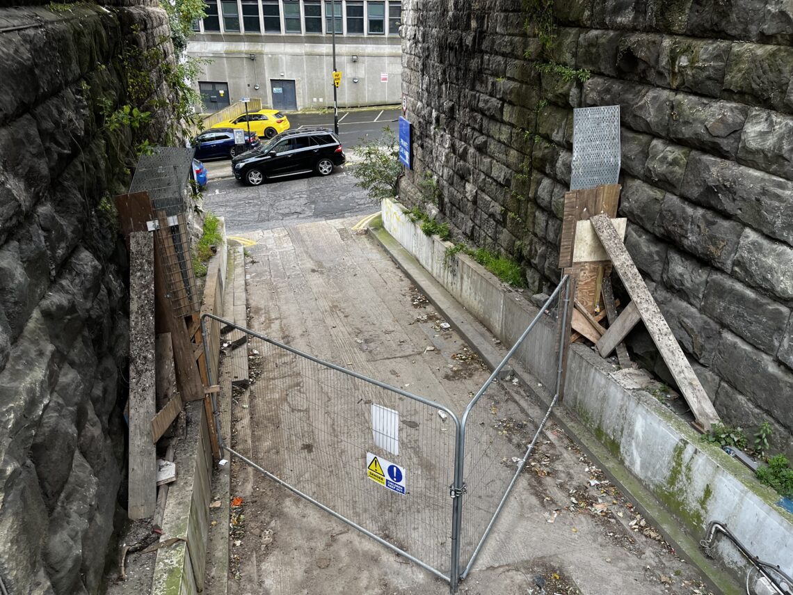 A blocked, sloping concrete path between stone walls leads to a city street. A metal fence and warning sign stop entry; wood planks and metal sheets lean on both sides. Across the street, a yellow taxi and black SUV are parked near a large gray building.