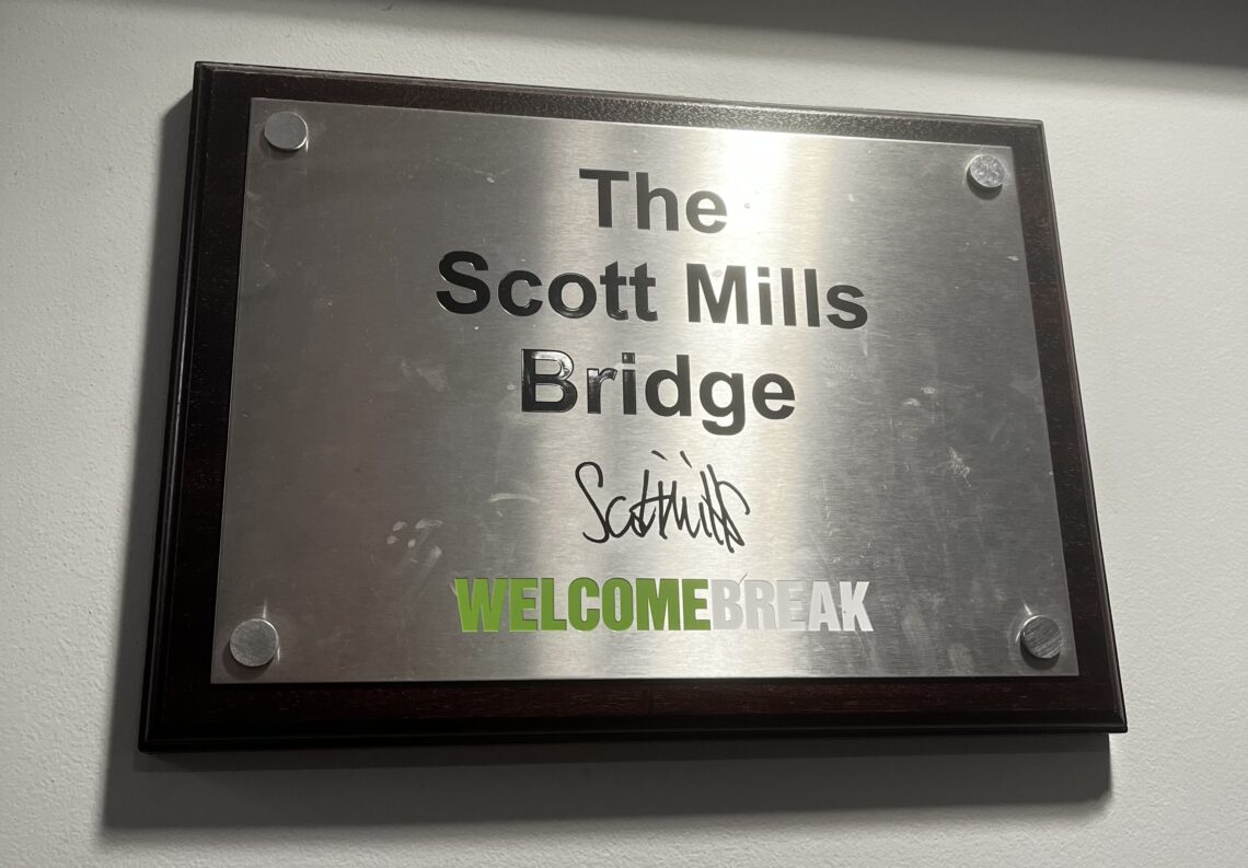 A brushed metal plaque in a dark wood frame reads The Scott Mills Bridge with Scott Mills signature below. At the bottom, WELCOMEBREAK is printed, with WELCOME in green and BREAK in white uppercase letters.