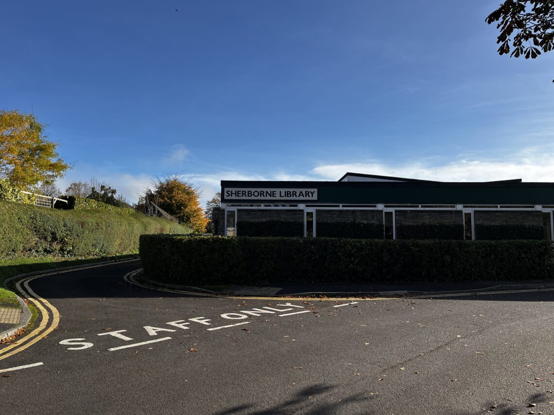 A single-story building with a sign reading “Sherborne Library” stands behind tall hedges. A road in the foreground has STAFF ONLY painted on it. The sky is clear and blue, with autumn trees and trimmed grass visible around the library.
