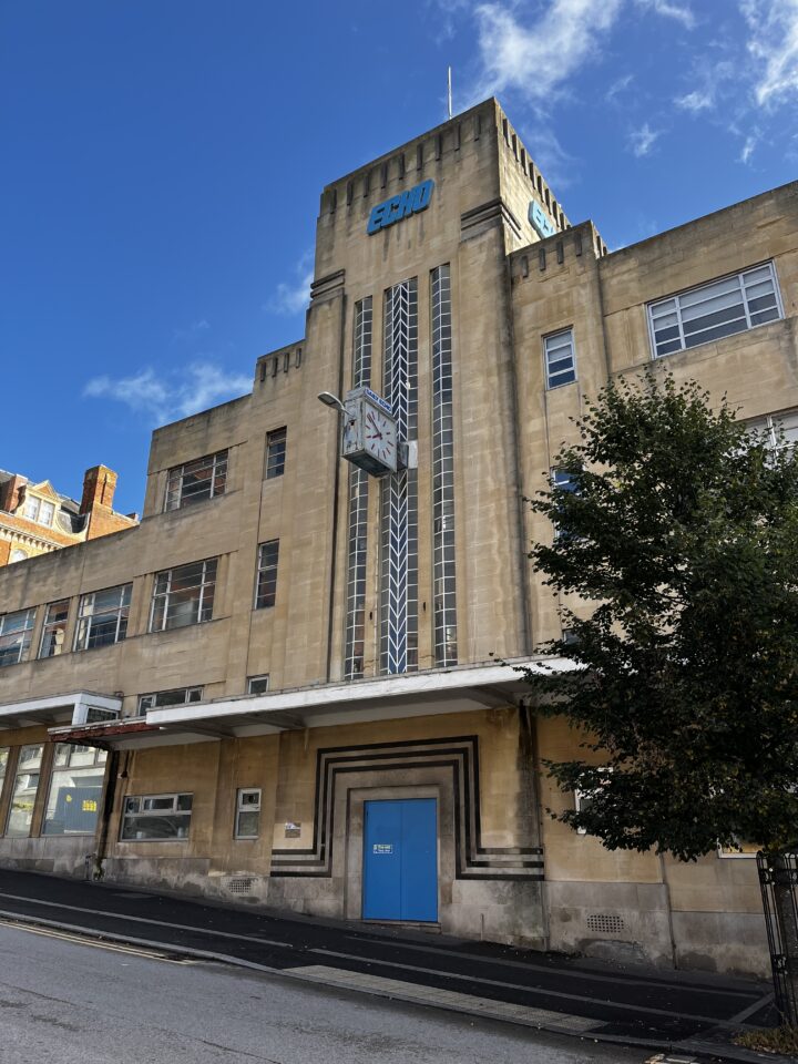 Art Deco building with tan stone façade, tall central tower, vertical windows, and geometric lines. Large clock projects above blue double doors. ECHO sign on top. Tree at right, blue sky with clouds above, and a sidewalk and quiet street in front.