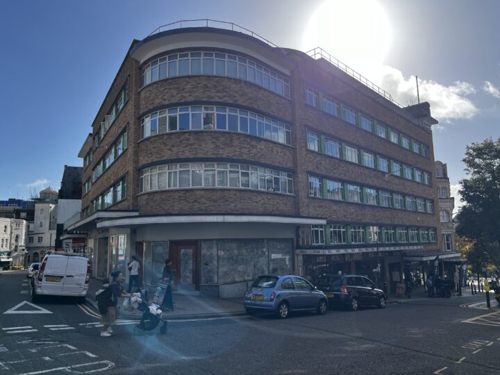 A curved corner brick building with many windows under a bright sun, casting shadows. Several cars are parked along the street, and a few people walk on the sidewalk, including a person pushing a stroller. The sky is mostly clear with a few clouds.