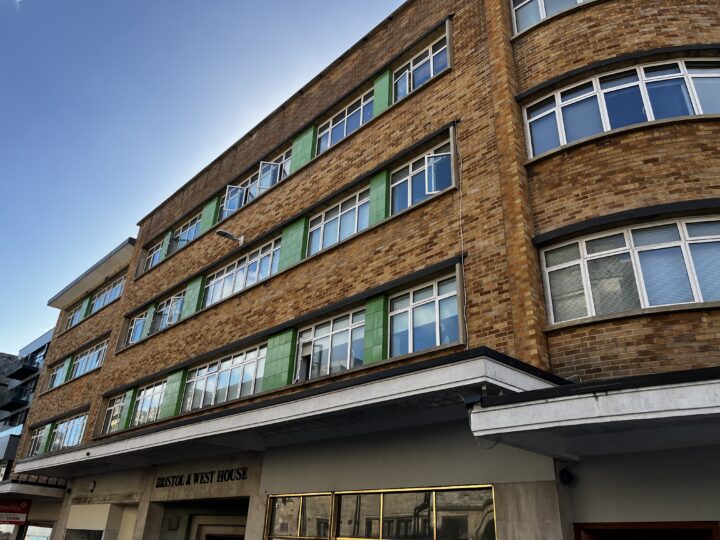 A four-story building features brown brick walls and large, rectangular windows with green vertical panels between some windows. A white awning extends above the entrance, labeled Bristol & West House, under a clear blue sky.
