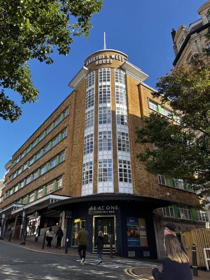 A tall, art deco building with Bristol & West House on the top, features curved brickwork, tall windows, and a Be At One cocktail bar on the ground floor. People walk nearby under a clear blue sky, with leafy trees framing the right side.