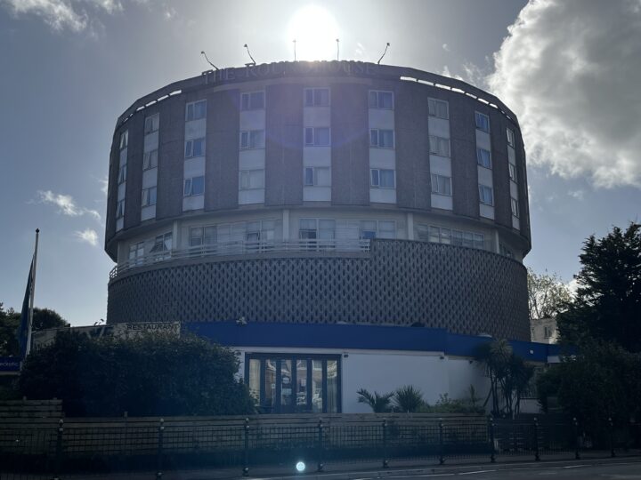 A round, modern hotel building with several floors and many windows, “The Roundhouse” sign above. The building is backlit by the sun, creating a halo effect. Blue sky and some clouds are visible; palm-like plants and a fence are in the foreground.