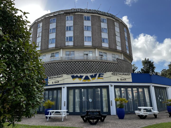 A round, multi-story gray building labeled “THE ROUNDHOUSE” sits above a blue and white restaurant called “WAVE RESTAURANT & BAR.” Patio tables, benches, plants, and trees are in the foreground on a sunny, partly cloudy day.