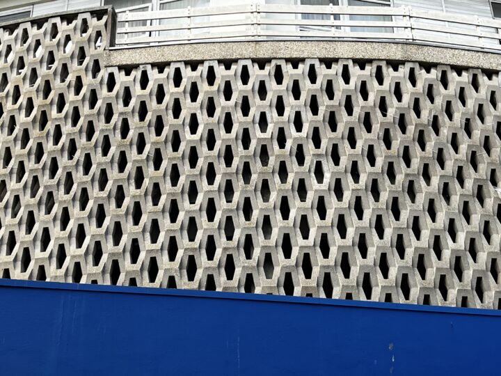 A wall with a geometric pattern of repeating hexagonal openings made from concrete is seen behind a solid blue construction barrier. Above the patterned wall, white railings and part of a window are visible. The angle is slightly upward.