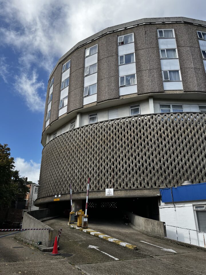 A multi-story building with patterned concrete facade and many windows rises above a sloped parking garage entrance. Two parking barriers and signs are at the entrance, with a traffic cone and tape nearby. Blue sky and clouds are visible overhead.