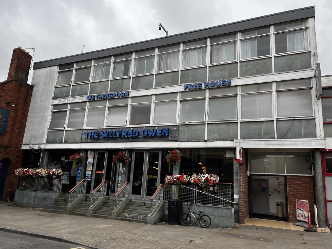A three-story, grey modernist building with large windows houses “The Wilfred Owen,” a Wetherspoon Free House pub. Flower baskets hang by the entrance, bikes are parked outside, and stairs lead to glass doors. The sky is overcast.