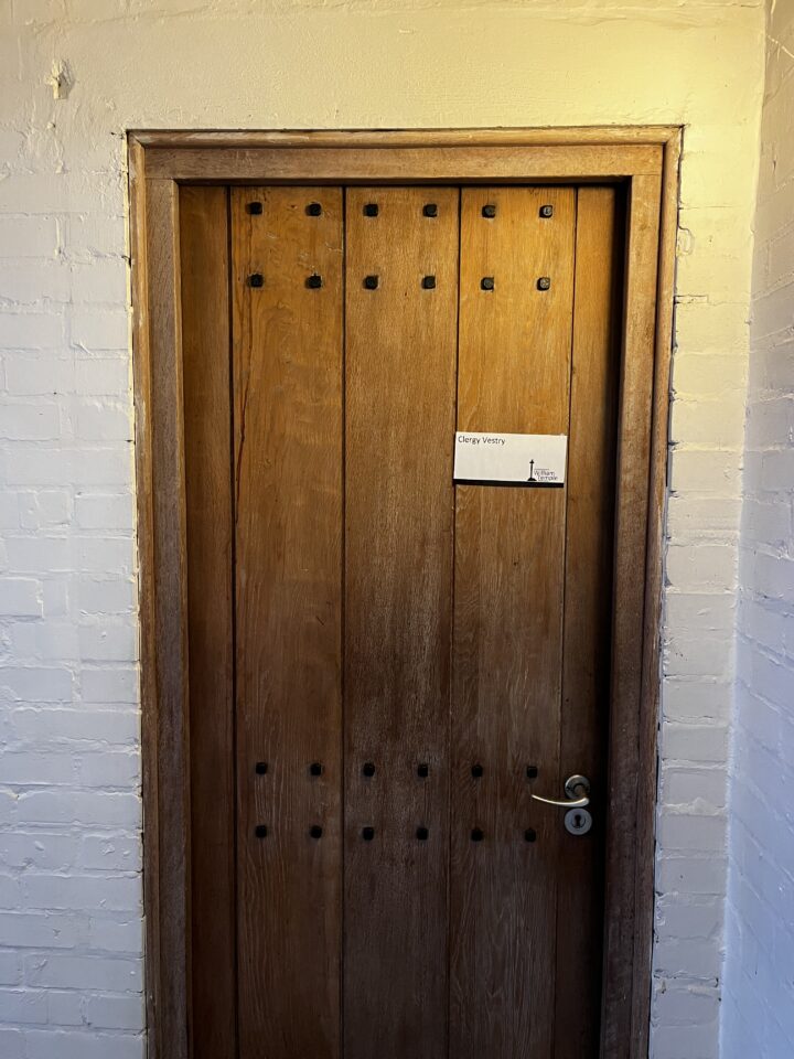 A solid wooden door with vertical panels and black studs is set in a white brick wall. There is a small sign on the door that says “Clergy vestry” with a simple illustration of a church on it. The door has a modern silver handle.