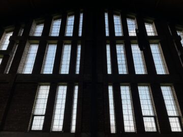 Large vertical windows with rectangular glass panes are set in a brick wall. The image is taken from inside, with daylight coming through, creating a strong contrast and making the interior appear dark and shadowed.