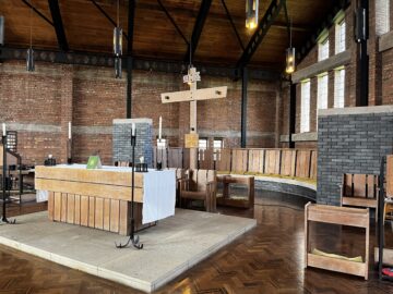 A modern church interior with red brick walls, wooden ceiling beams, and tall windows. The central altar features a white cloth and wooden elements. Wooden benches and a large cross hang above, with soft natural light illuminating the space.