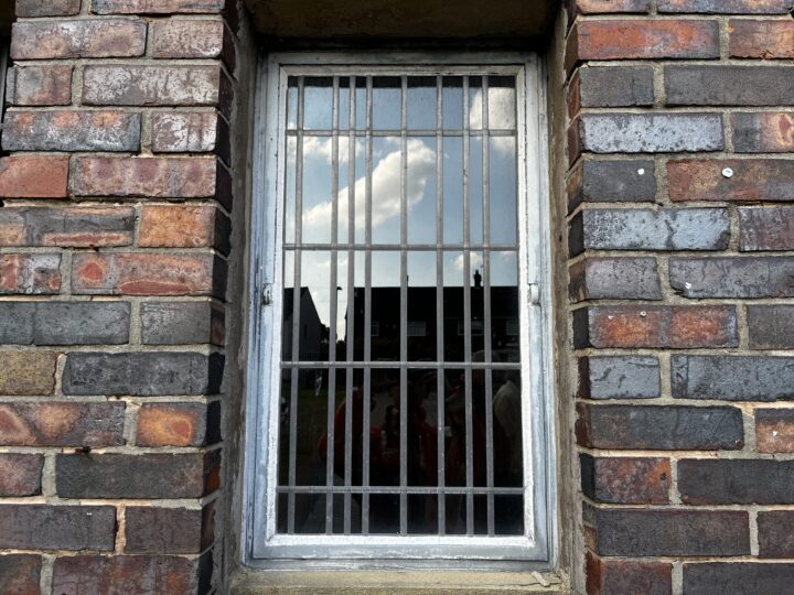 A rectangular window with vertical and horizontal metal bars is set in a wall of multicolored, weathered bricks. The glass reflects a cloudy sky and faint outlines of nearby buildings and people.