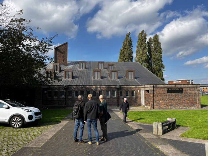 Four men stand on a paved path leading to a brick building with a sloped roof and multiple dormer windows under a blue sky with clouds. A white car is parked on the left and trees and grass surround the area. One man walks toward the entrance.