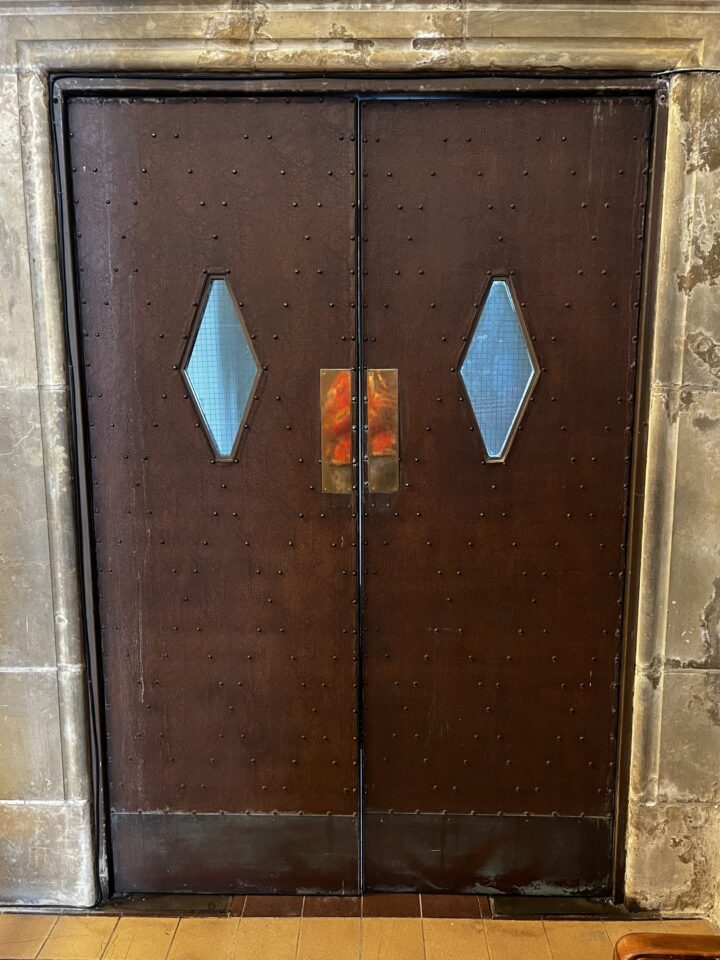 A pair of dark brown, textured metal double doors with small diamond-shaped windows, set in a weathered stone or concrete frame. The doors have a central rectangular metal push plate, and rivets are visible along the door surface.