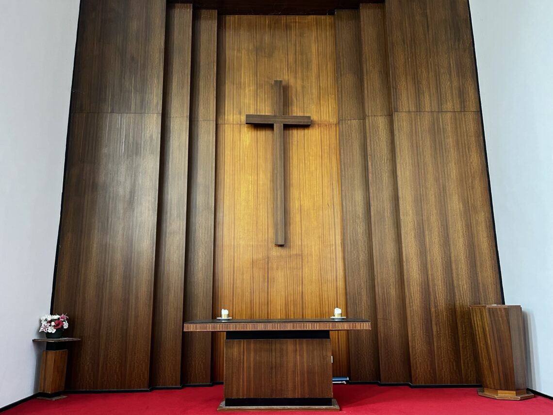 A church altar against a tall wooden-paneled wall with a large wooden cross in the center. The altar has two white candles and a cross, with a flower arrangement on the left and a wooden podium on the right. The floor is covered in red carpet.